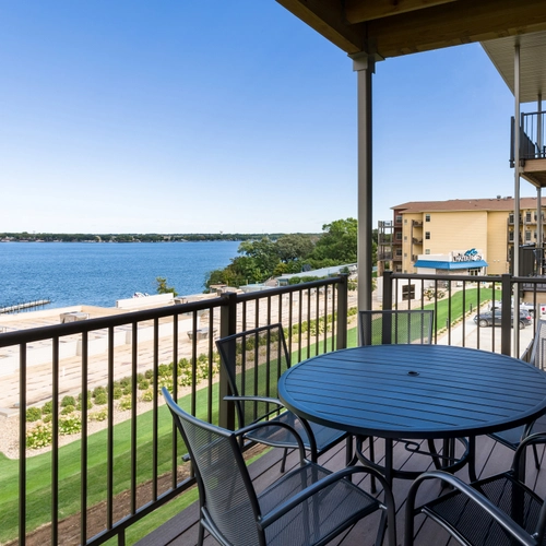 A round table with four chairs sits on a balcony overlooking a lake, with a clear blue sky, grassy area, and nearby buildings visible in the background.