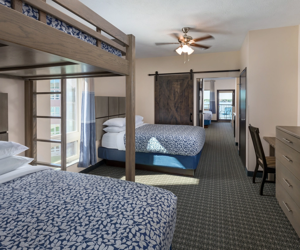 Hotel room with a bunk bed, a double bed, a dresser with a TV, a desk with a chair, sliding barn door, and carpeted floor. Large window lets in natural light.