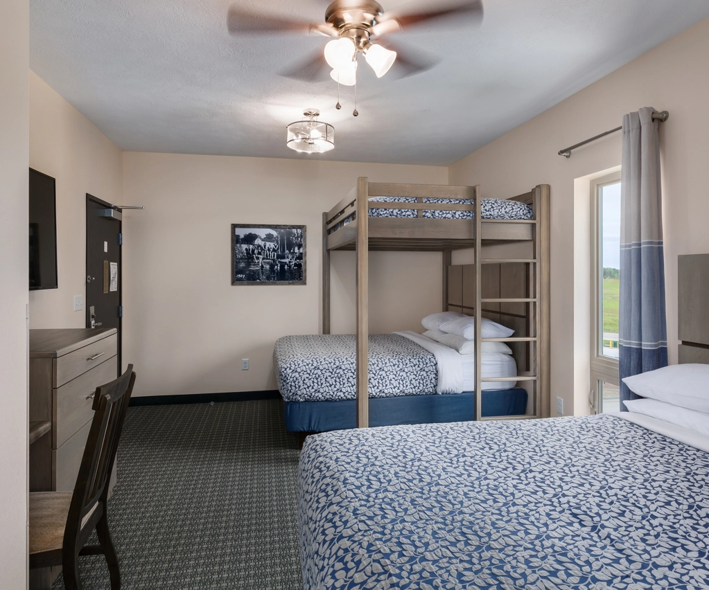 Hotel room with a double bed, bunk bed, desk with chair, wall-mounted TV, a window with curtains, and a ceiling fan. The room has neutral walls and patterned bedding.