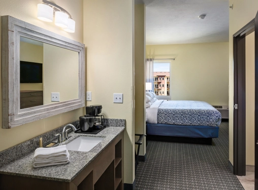 A hotel room with a granite vanity sink, towels, coffee maker, and mirror in the foreground. In the background, there is a bed with white and blue linens near a window.