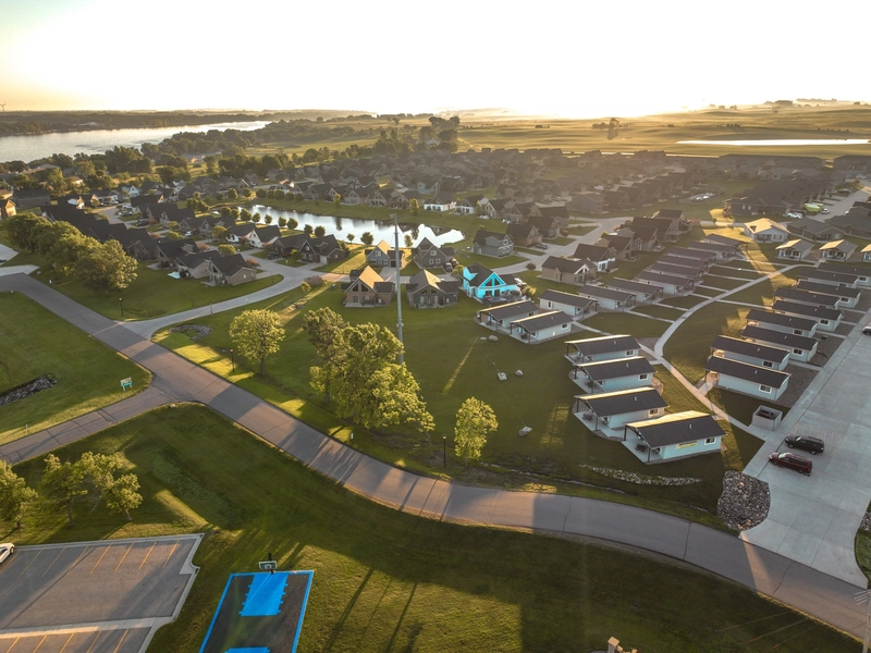 Aerial view of a suburban neighborhood at sunset with houses, green lawns, winding roads, ponds, and trees, and a large open field. Some homes have light blue highlights on roofs and driveways.