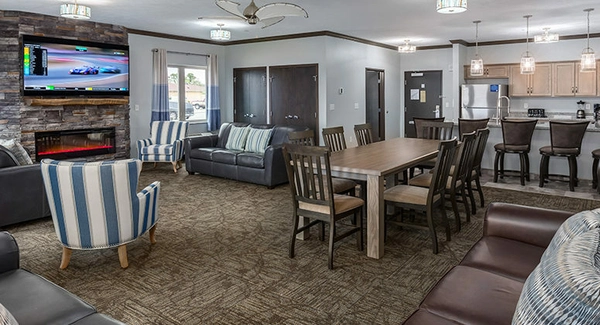 Modern living room with sofas, armchairs, a dining table, and a kitchen with barstools. TV above a fireplace shows a race. Pendant lights hang over the kitchen counter. Neutral tones with blue accents throughout.