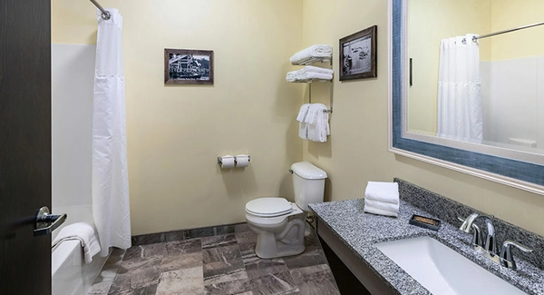 A clean hotel bathroom with a granite countertop, sink, toilet, and bathtub with a white shower curtain. Towels are neatly stacked on shelves, and framed pictures hang on the light yellow walls.