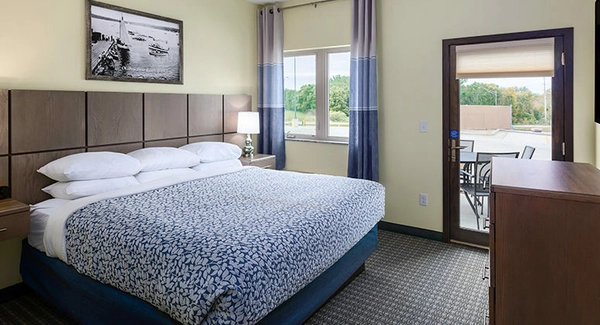 A hotel room with a king bed, blue and white bedding, two nightstands with lamps, a window with curtains, a door leading to a patio, a dresser, and a black and white photo above the bed.