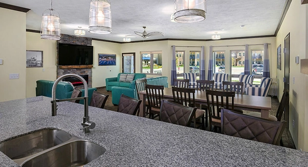 Large open-concept living and dining area with granite kitchen island, dining table, teal sofas, striped armchairs, wall-mounted TV, and large windows letting in natural light.
