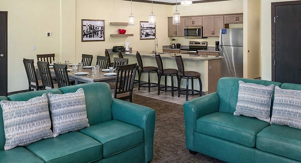 Modern open-concept living space with two teal sofas, a dining table with eight chairs, a kitchen with barstools, stainless steel appliances, pendant lights, and framed photos on light yellow walls.