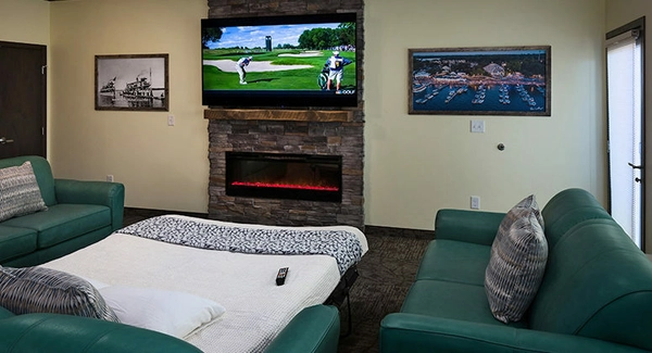 A cozy living room with teal sofas, a pull-out bed, a TV above a stone fireplace showing golf, wall art, and a remote on the bed. Natural light comes in from a window and glass door.