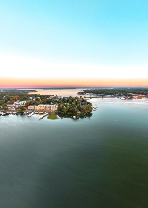 An aerial view at sunset shows a tranquil lake with a peninsula extending into the water, dotted with buildings, dense trees, and numerous docks. The horizon glows with soft orange and pink hues, reflecting onto the calm surface of the water.