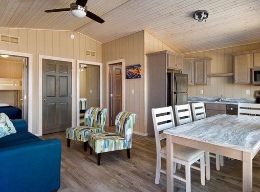 A cozy cabin interior with wood walls, open kitchen, dining table, blue sofa, two patterned chairs, and visible bedroom with bunk beds. Ceiling fan and modern appliances are present.