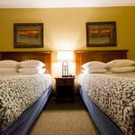 Two double beds with patterned bedspreads and multiple pillows, separated by a nightstand with a lit lamp. Two framed pictures hang above the beds on a beige wall.