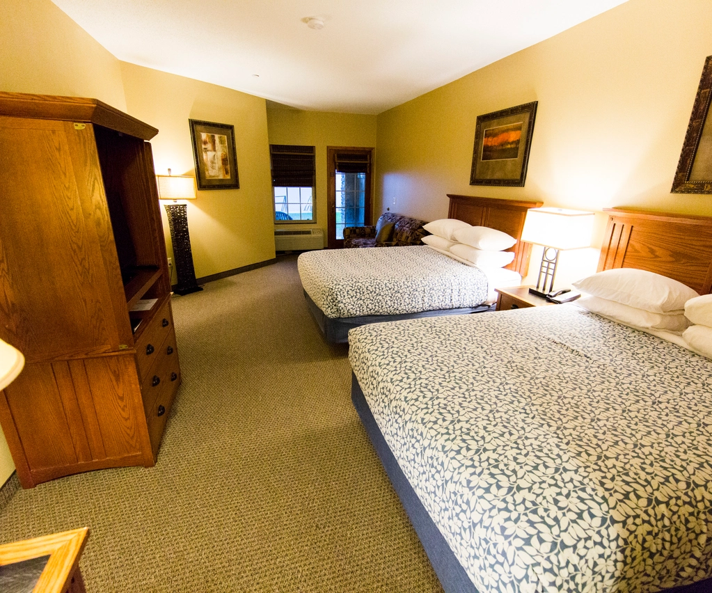 Hotel room with two queen beds, wooden headboards, a dresser with TV, lamps, framed art on beige walls, and a window with brown blinds in the back.