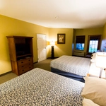 A hotel room with two double beds, a wooden TV cabinet, a lamp, framed art on the walls, beige walls, and a window with a view of trees outside.