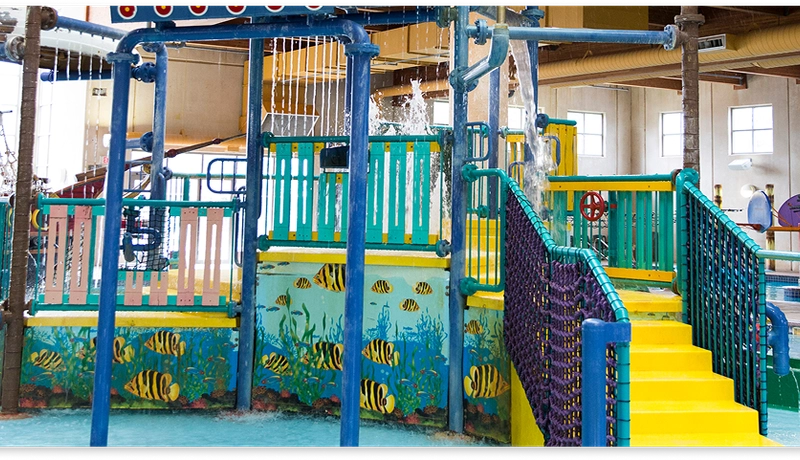 Indoor water play area with colorful climbing structures, yellow stairs, nets, and painted fish. There’s a pirate ship in the background and a pool with water jets nearby. Bright lighting illuminates the space.